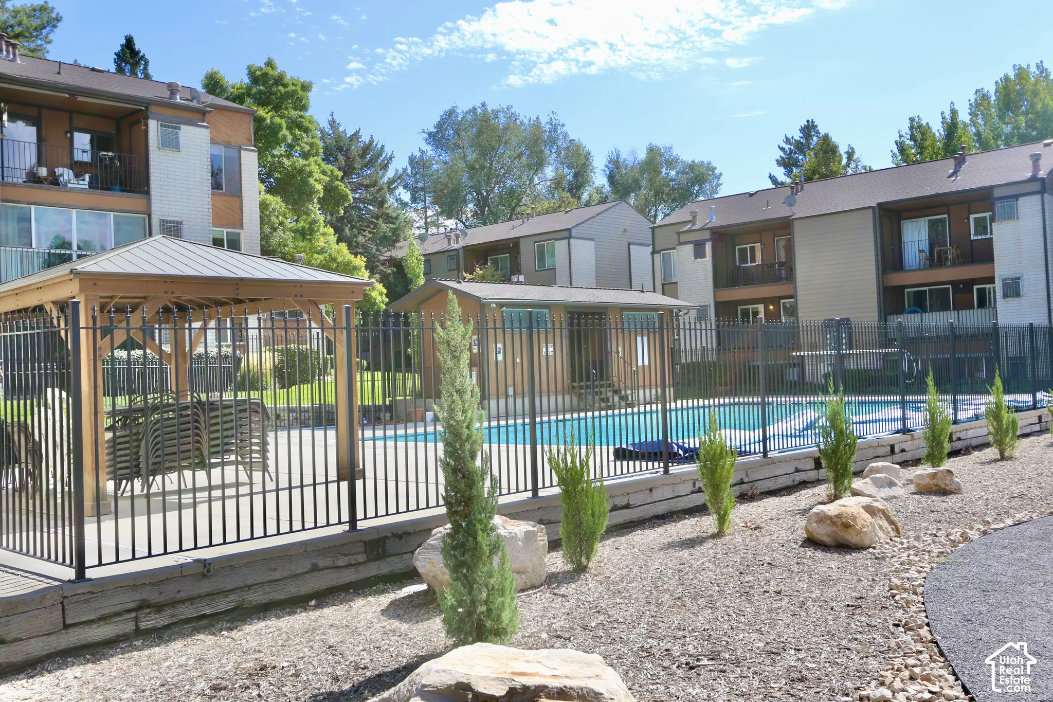 Outdoor swimming pool and clubhouse with black metal fencing, adjacent wooden gazebo, and surrounding gravel landscaping with decorative rocks and small evergreen plantings