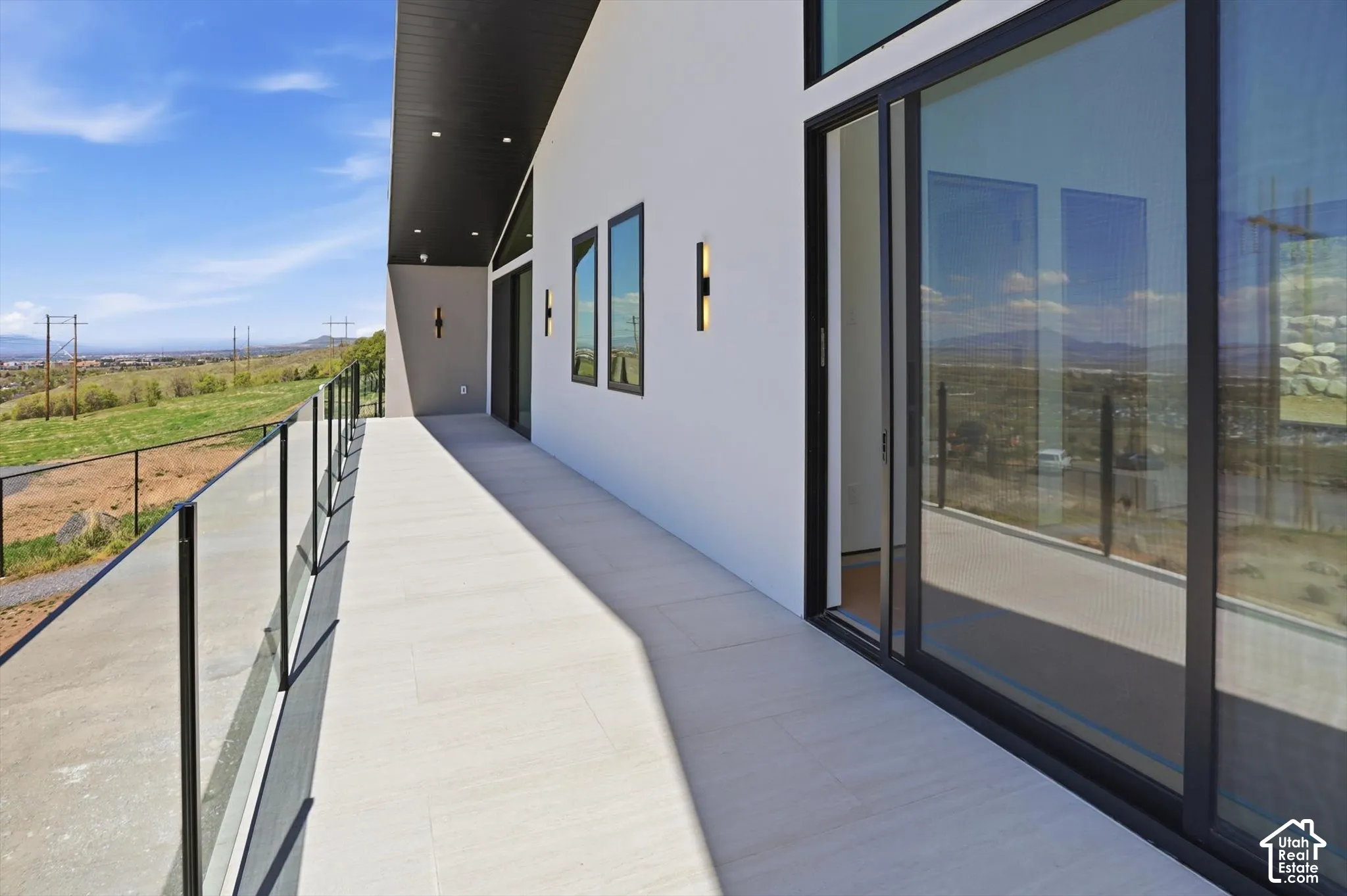 Expansive exterior balcony with glass railing, contemporary wall sconces, recessed ceiling lighting, and large sliding glass doors