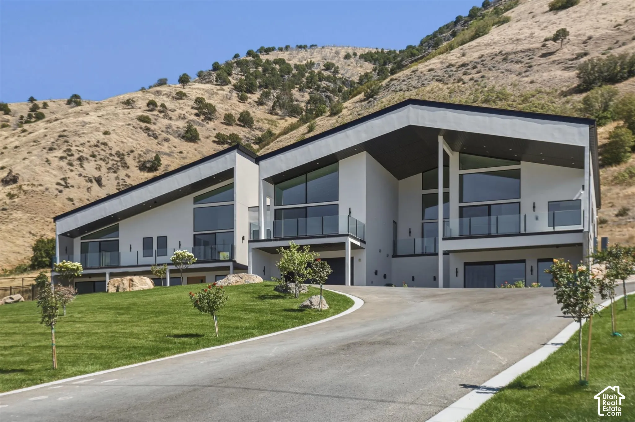 Modern architectural design with a sloped roofline, prominent balconies featuring glass railings, a paved driveway, and a manicured lawn