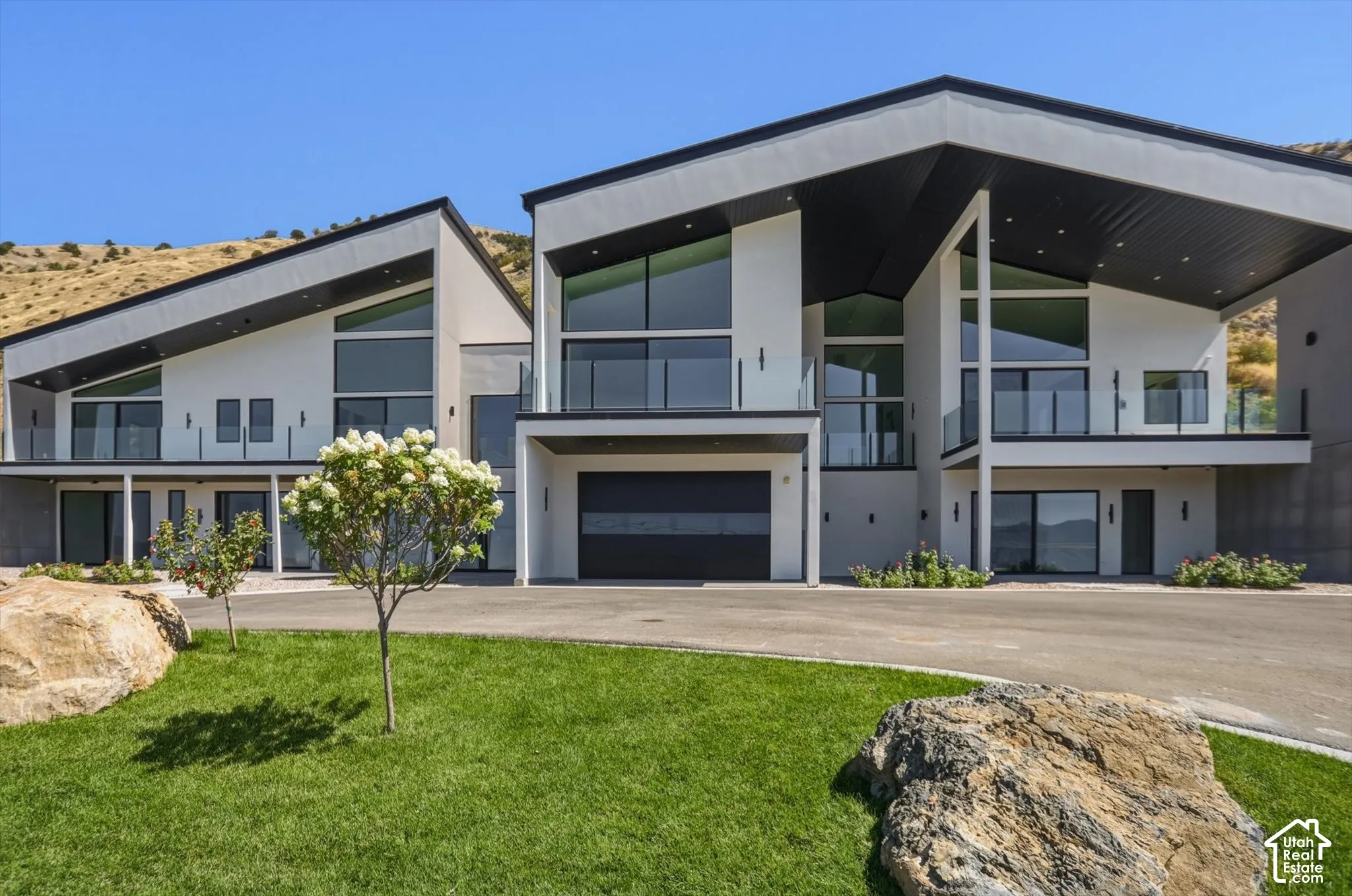 Modern architectural design featuring expansive glass panels, multiple balconies with glass railings, two integrated garages with contemporary doors, and a sloped roofline