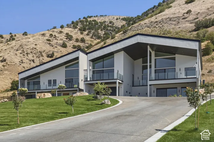 Modern architectural design with a sloped roofline, prominent balconies featuring glass railings, a paved driveway, and a manicured lawn