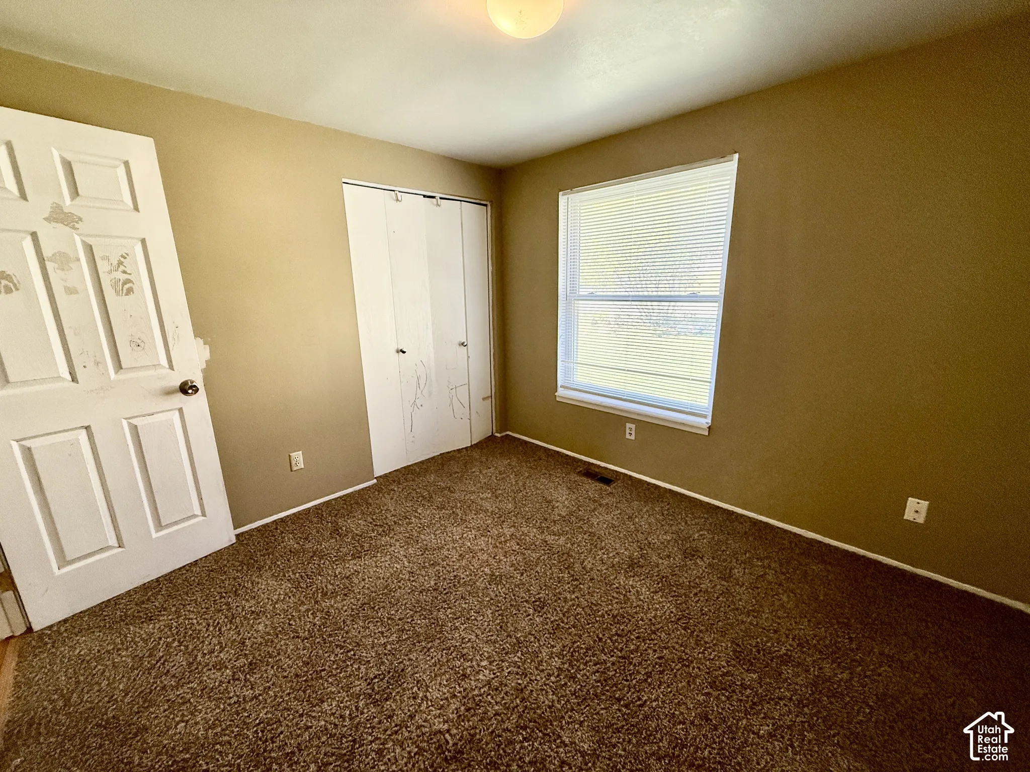 Carpeted room featuring a single window with blinds and white trim, and a bi-fold closet door