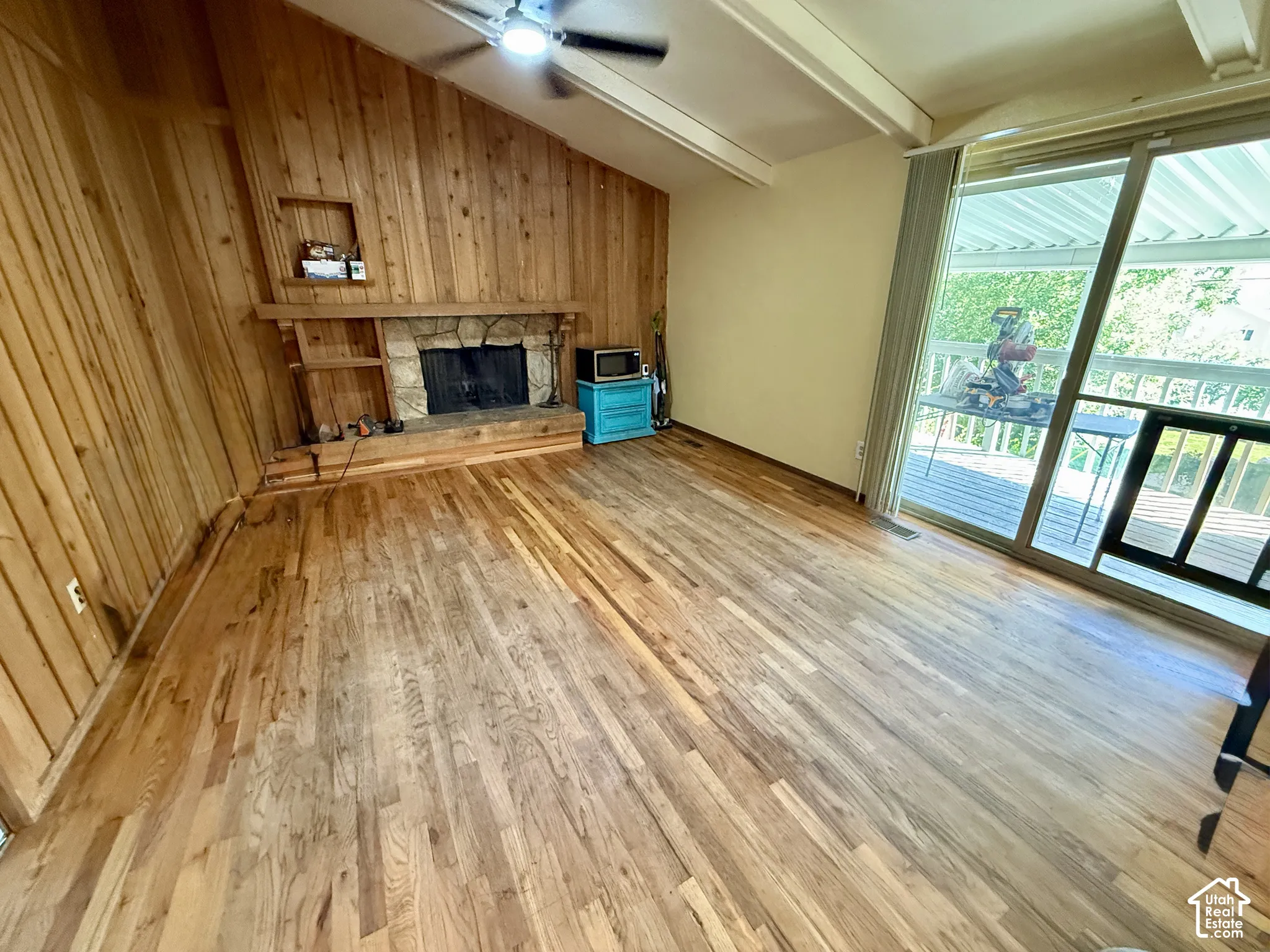 Hardwood flooring, wood-paneled accent wall, stone-clad fireplace with built-in shelving, vaulted ceiling with exposed beams, and sliding glass door