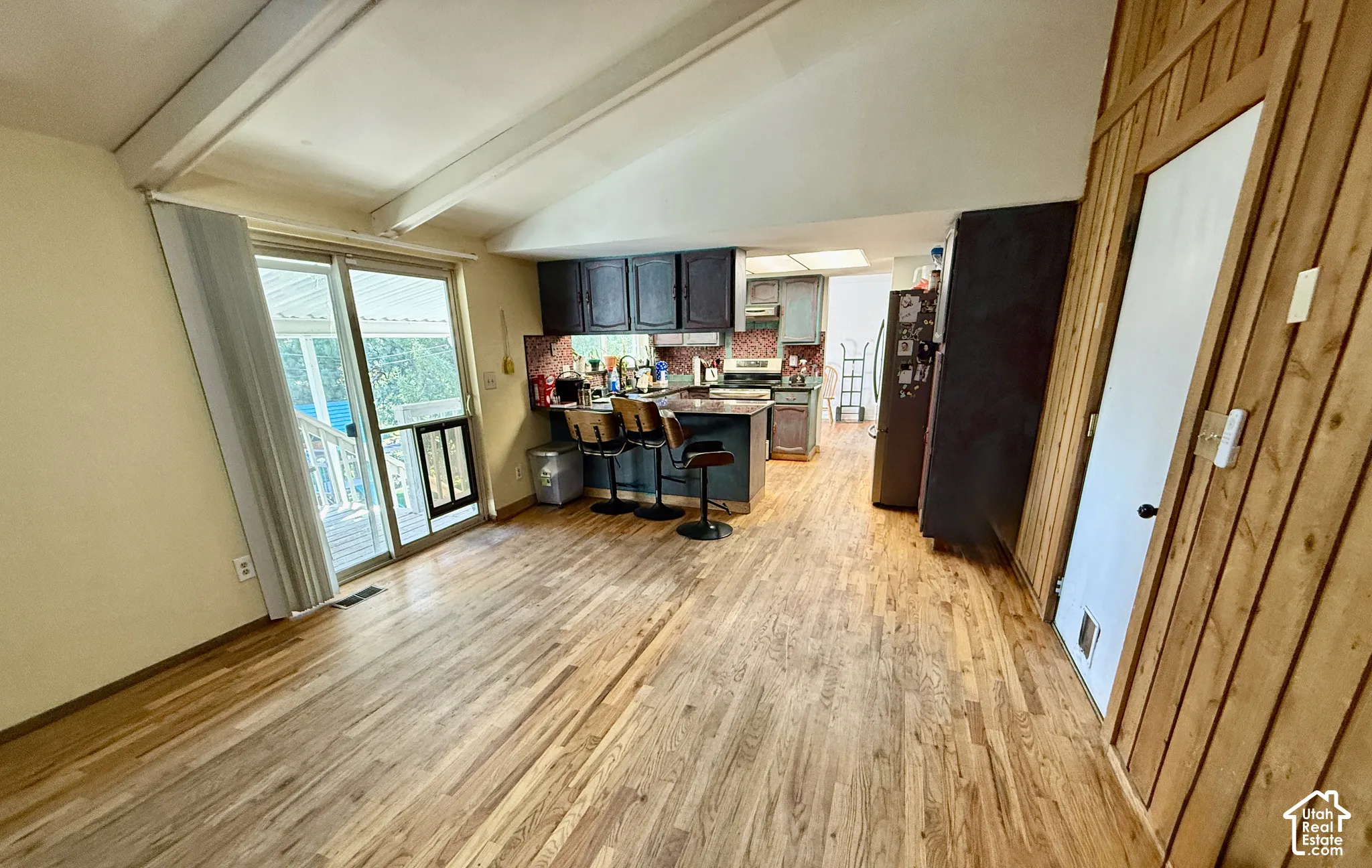 Vaulted ceilings with exposed beams, wood-finish flooring, sliding glass door, kitchen with dark cabinetry, and a central island with bar seating
