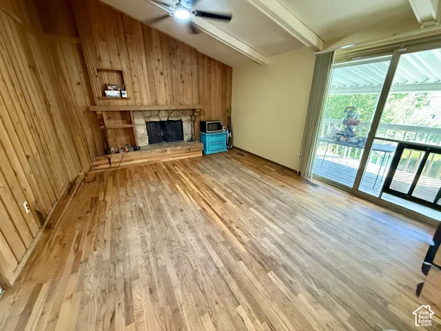 Hardwood flooring, wood-paneled accent wall, stone-clad fireplace with built-in shelving, vaulted ceiling with exposed beams, and sliding glass door