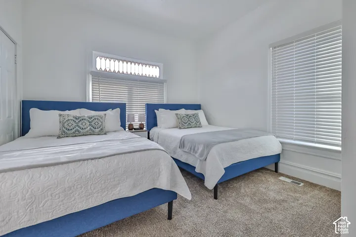 Bright guest bedroom suite featuring two beds with navy upholstered headboards, crisp white bedding with decorative pillows, warm carpet flooring, and abundant natural light from dual windows with horizontal blinds