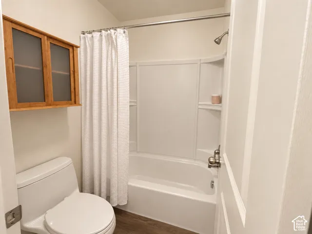 White toilet, wood-finish flooring, built-in wall cabinet with frosted glass panels, white bathtub with integrated shelving, and a chrome shower fixture