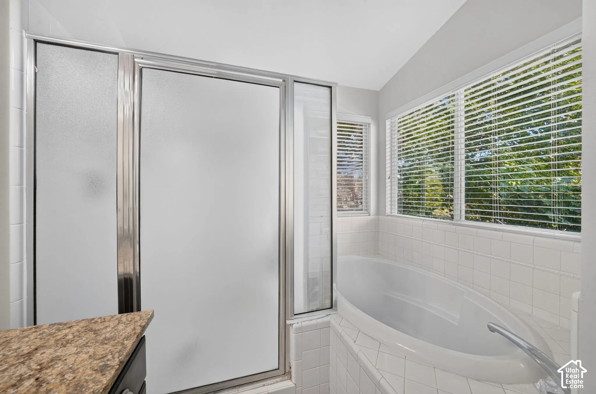 Spa-inspired bathroom featuring a curved soaking tub, frameless glass shower enclosure with frosted panels, white subway tile surround, quartz countertop with integrated sink, and abundant natural light from multi-pane windows with plantation shutters