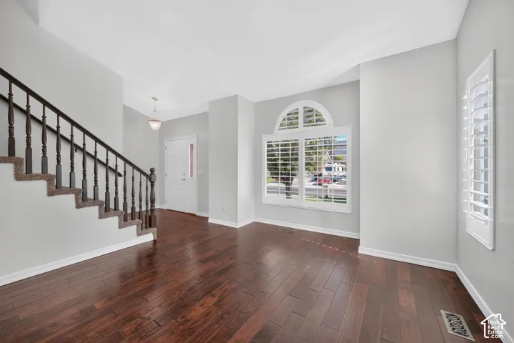 Hardwood-finish flooring, staircase with carpeted treads and dark wood balusters, arched top window with plantation shutters, additional window with plantation shutters, and a front entry door with a decorative glass panel