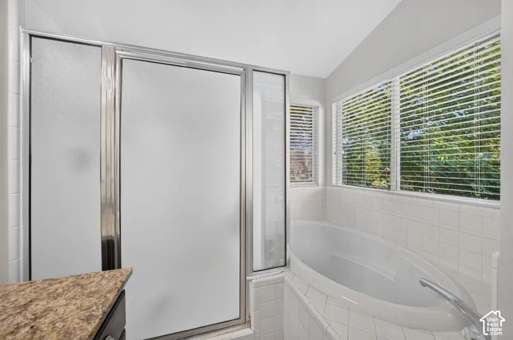 Spa-inspired bathroom featuring a curved soaking tub, frameless glass shower enclosure with frosted panels, white subway tile surround, quartz countertop with integrated sink, and abundant natural light from multi-pane windows with plantation shutters