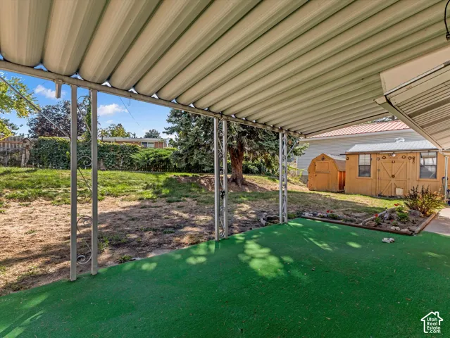 Covered patio with green artificial turf, supported by ornate metal columns, leading to a backyard with a storage shed and mature trees