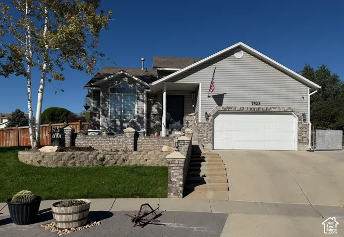 Multi-level residence featuring a two-car garage, stone and siding exterior, large concrete driveway, and tiered landscaping with mature trees