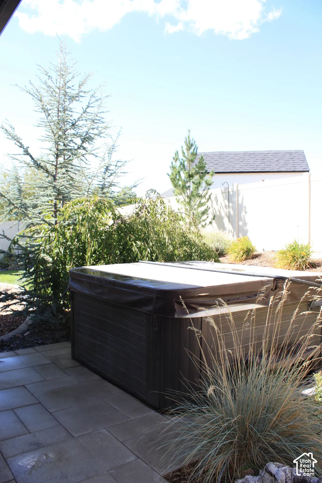 Paved patio area featuring a covered spa, surrounding evergreen trees, and integrated landscaping with ornamental grasses