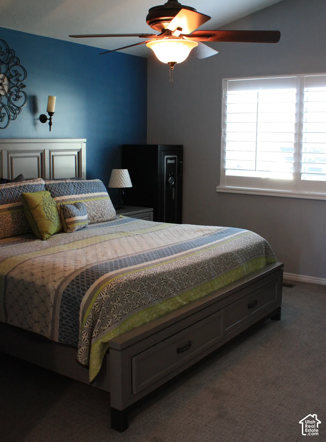 Vaulted ceiling, ceiling fan with light fixture, window with plantation shutters, wall-mounted sconce, and carpet flooring