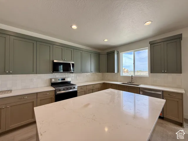 Modern kitchen featuring a large quartz-topped island with waterfall edge, stainless steel appliances including a gas range, white subway tile backsplash, soft-close cabinetry in sage and warm wood tones, recessed lighting, and abundant natural light from