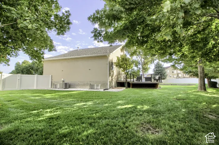 Expansive green lawn with mature trees, stucco exterior, rear deck with dark railings, and a light-colored privacy fence
