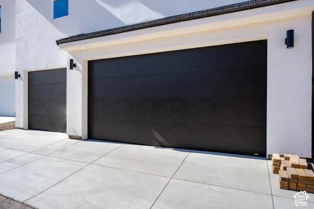 Smooth-finish black garage doors, white stucco exterior, black wall-mounted sconce lighting, and a concrete driveway