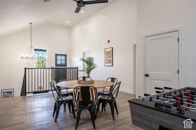 Open-concept dining and recreation area featuring a round wood-finish table, black metal chairs, wide-plank wood-finish flooring, soaring vaulted ceilings with ceiling fan, and abundant natural light from upper windows