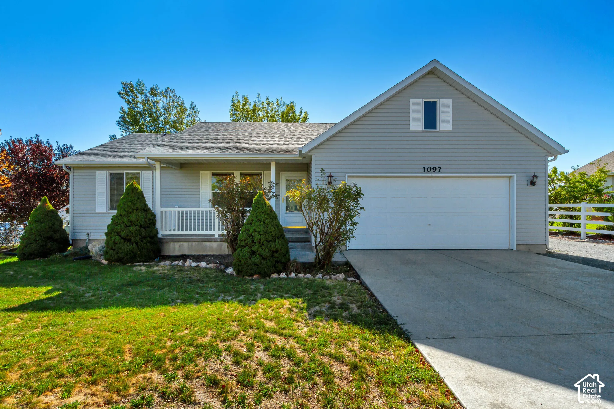 Charming ranch-style home featuring a front-facing two-car garage, covered front porch with decorative railings, and manicured landscaping with mature evergreen plantings