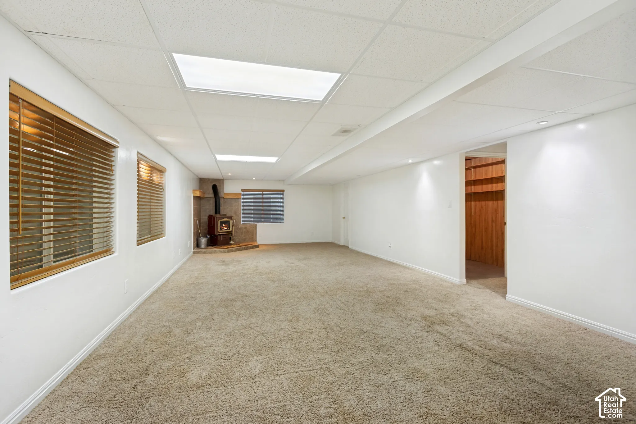 Spacious room with plush carpeting, wood-burning stove with stone surround, recessed lighting, and drop ceiling with integrated lighting
