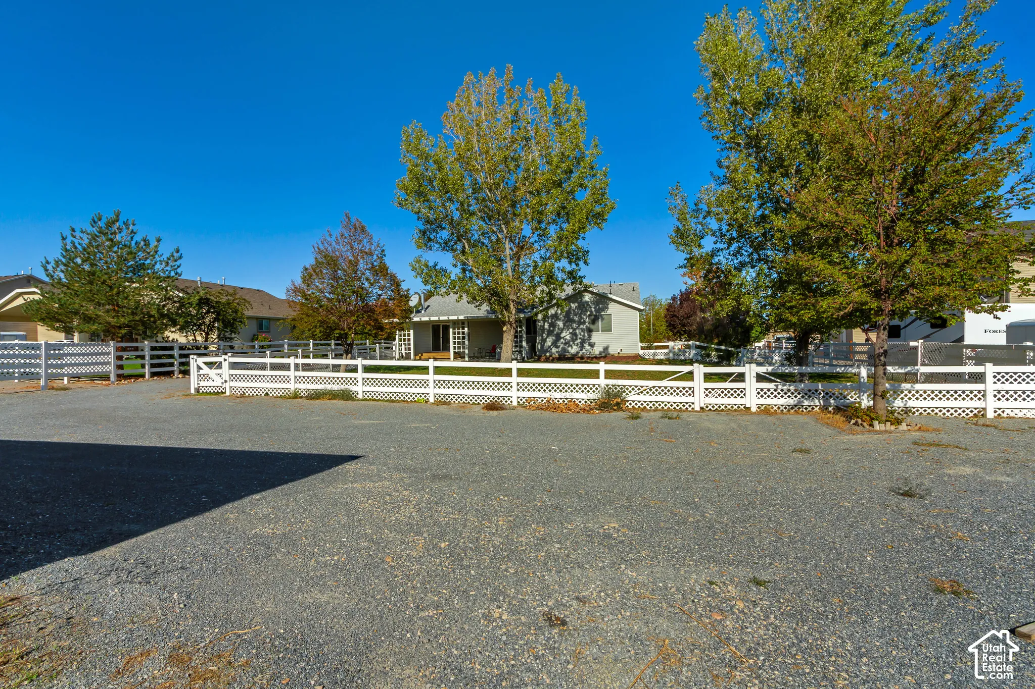 Expansive gravel parking area with a white lattice fence and gate, featuring mature trees and a distant single-story structure