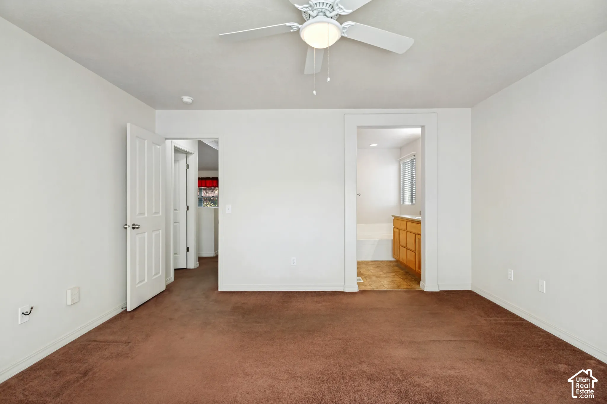 Brown carpeted flooring, white walls, ceiling fan with integrated lighting, white interior doors, and wood-finish cabinetry visible in the background