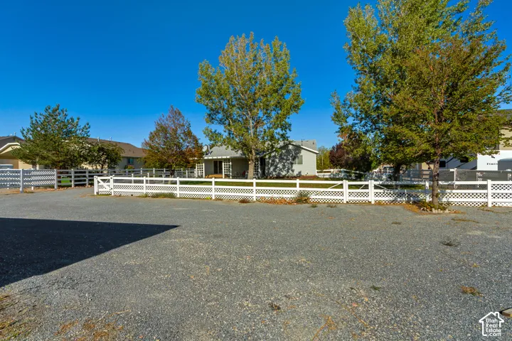 Expansive gravel parking area with a white lattice fence and gate, featuring mature trees and a distant single-story structure