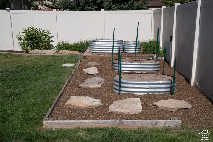 Backyard garden area with four galvanized raised beds, rock-lined pathway, wood chip ground cover, and a white privacy fence