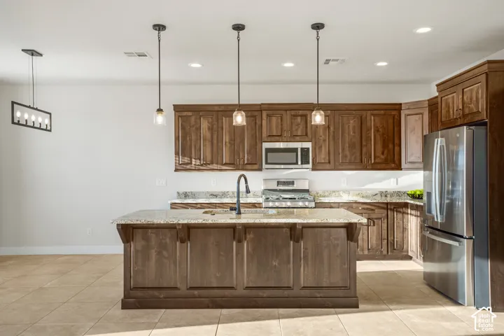 Kitchen featuring wood-finish cabinetry, stainless steel appliances, granite countertops, a central island with a sink, and tile flooring
