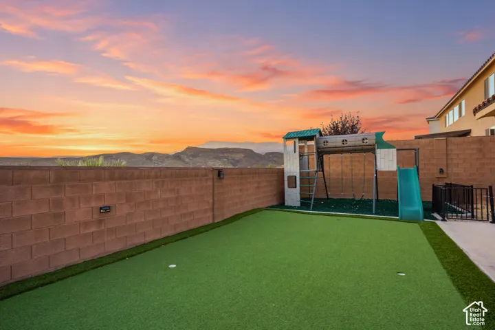 Artificial turf putting green with two holes, concrete block perimeter fencing, and a built-in play structure featuring a slide and swings