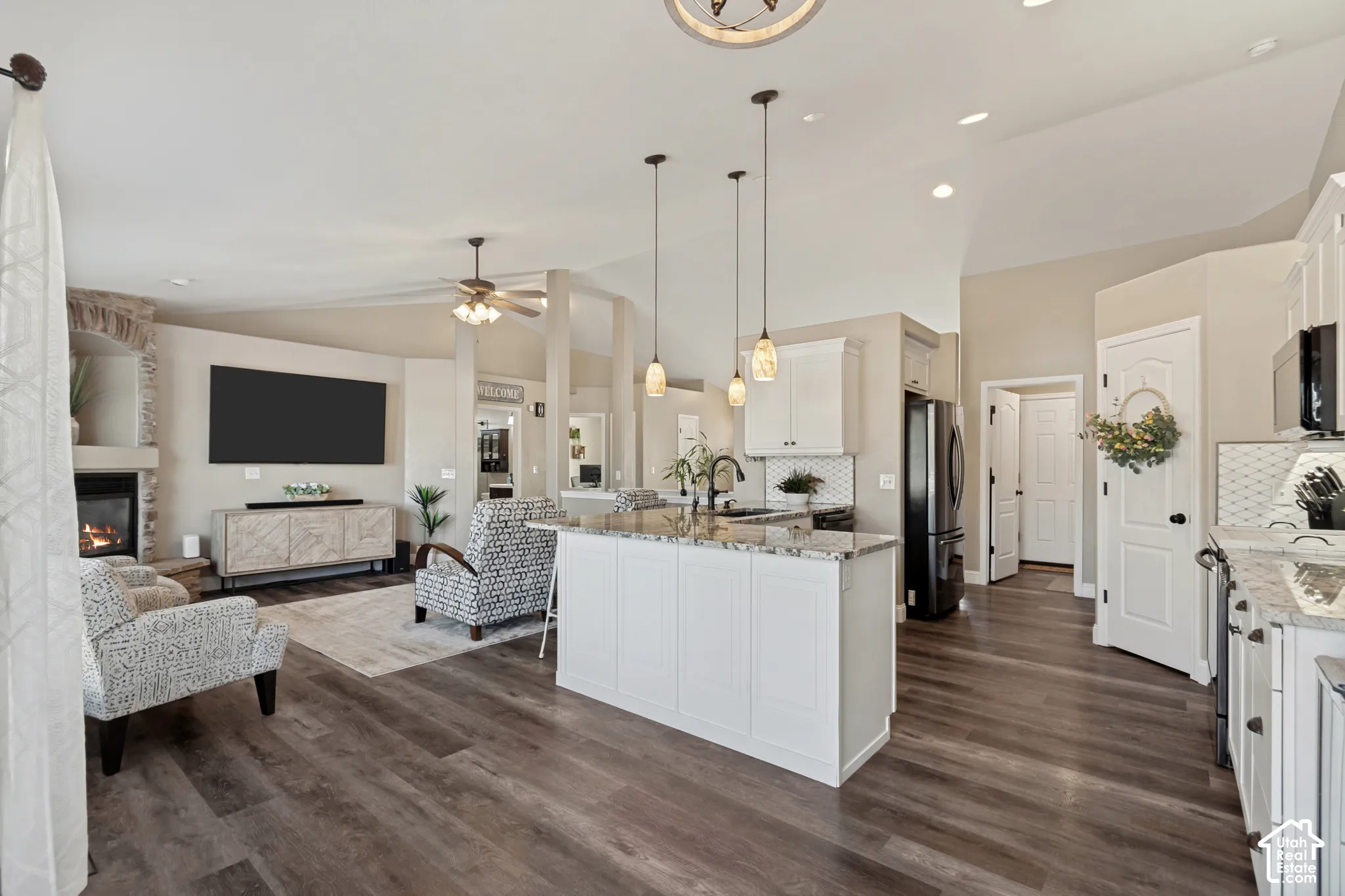 Open-concept living space featuring wood-finish flooring, a stone-surround fireplace, vaulted ceilings, a kitchen island with pendant lighting, and white cabinetry