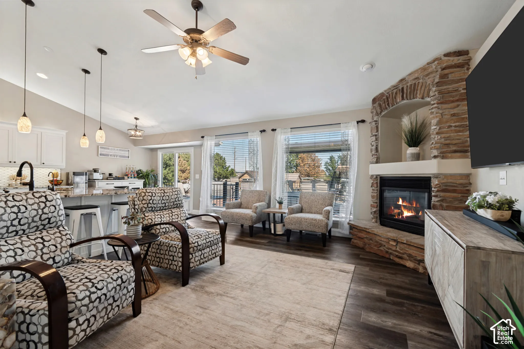Open-concept living area with wood-finish flooring, a stone-faced fireplace with hearth, and a ceiling fan