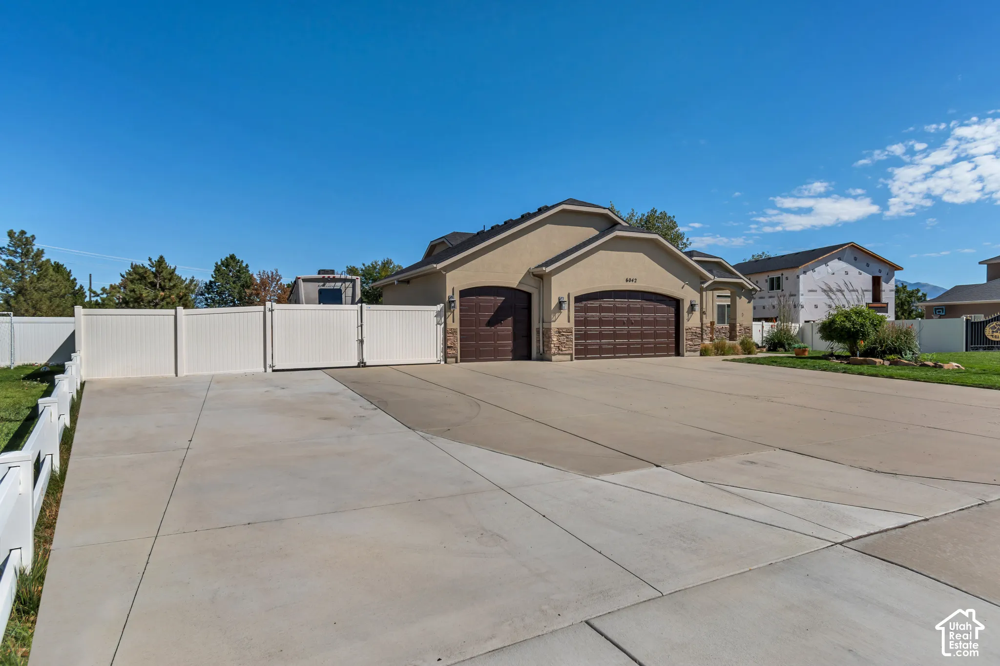 Stucco exterior with stone accents, two attached garages with arched doors, expansive paved driveway, white privacy fence, and manicured front landscaping