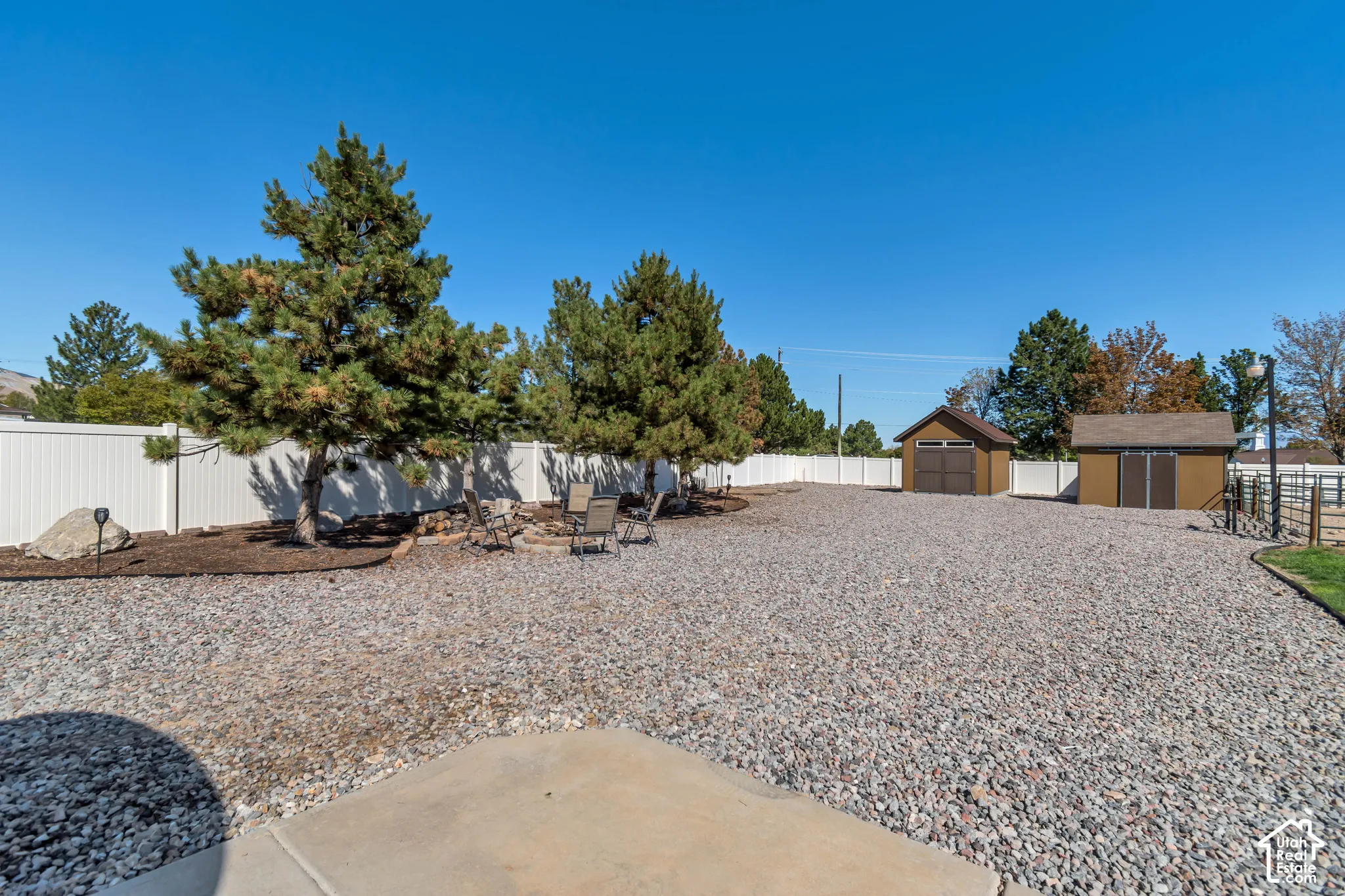 Expansive gravel yard featuring mature trees, a white perimeter fence, and two detached storage sheds