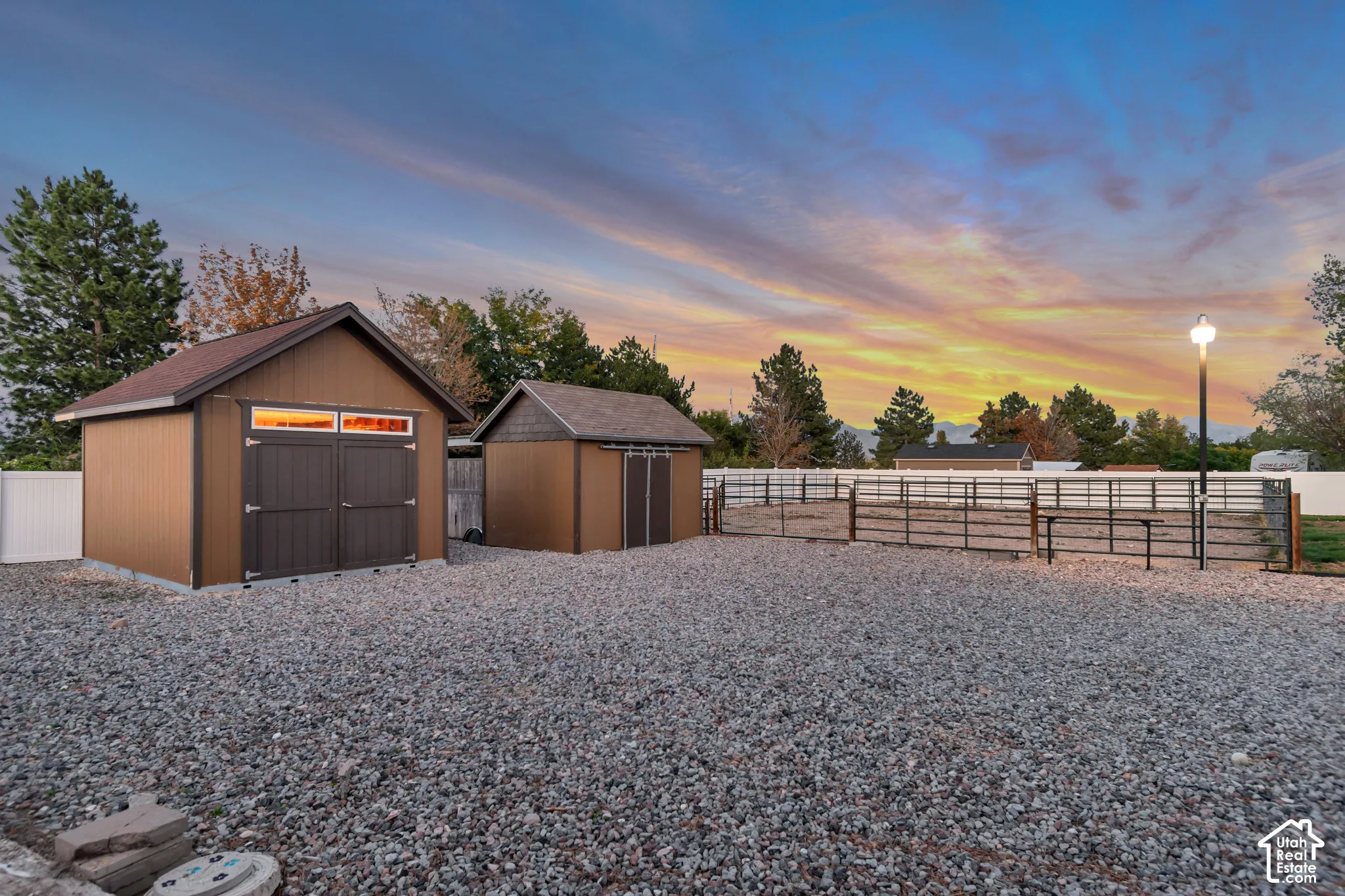 Gravel-covered outdoor area featuring two storage sheds, a white privacy fence, metal fencing, and a street light
