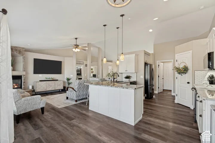 Open-concept living space featuring wood-finish flooring, a stone-surround fireplace, vaulted ceilings, a kitchen island with pendant lighting, and white cabinetry