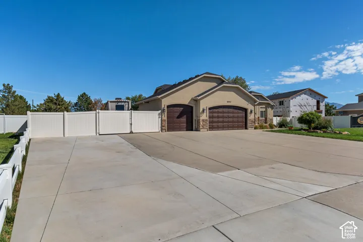 Stucco exterior with stone accents, two attached garages with arched doors, expansive paved driveway, white privacy fence, and manicured front landscaping