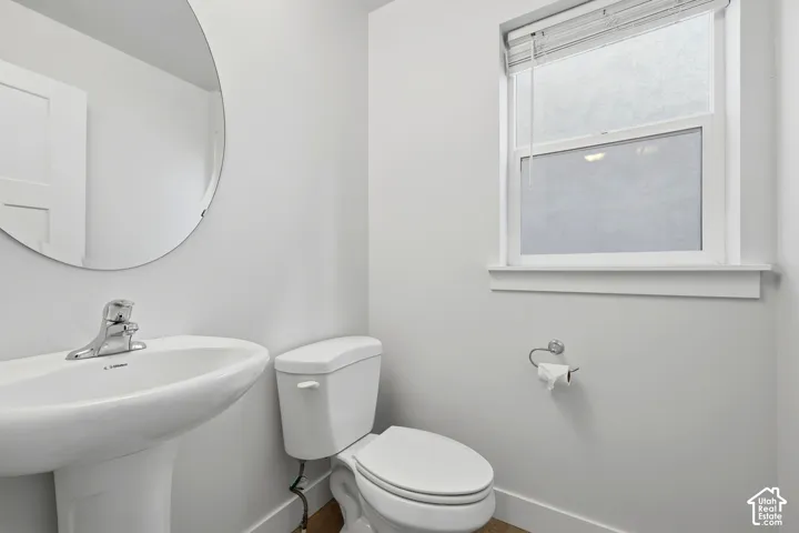 Pedestal sink with chrome faucet, circular wall mirror, window with privacy film, white toilet, and wood-finish flooring