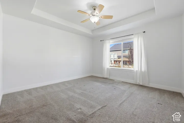 Tray ceiling with a ceiling fan, large window, and light-toned carpeting