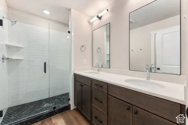 Dual vanity with integrated sinks, wood-finish flooring, frameless glass shower enclosure, black penny tile shower pan, and white subway tile shower surround