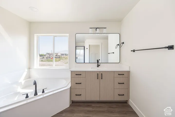 Corner soaking tub with matte black fixtures, wood-finish vanity with a light-toned countertop, rectangular framed mirror, and wood-finish flooring