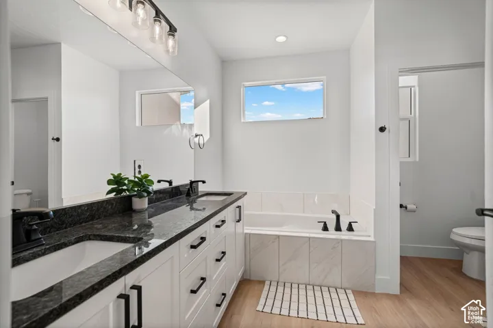 Double vanity with dark countertops and white cabinetry, built-in bathtub with tiled surround, matte black fixtures, and wood-finish flooring