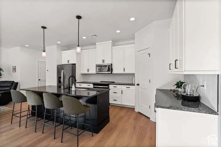 Kitchen featuring wood-finish flooring, white cabinetry with black hardware, an integrated refrigerator, a large center island with pendant lighting, and dark countertops