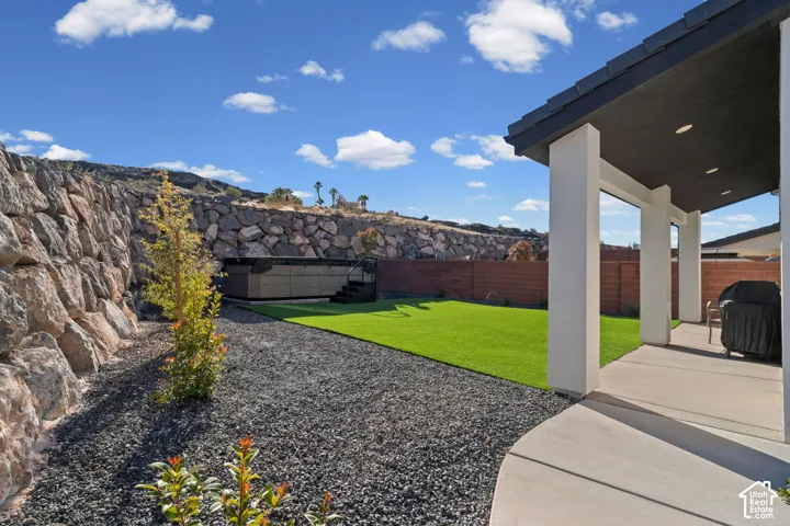 Covered patio with recessed lighting, integrated hot tub, artificial turf lawn, dark gravel landscaping, and a natural stone retaining wall