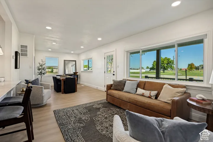 Bright, open-concept living space featuring expansive multi-panel windows framing pastoral landscape vistas, warm wood-finish flooring throughout, and a generous seating area