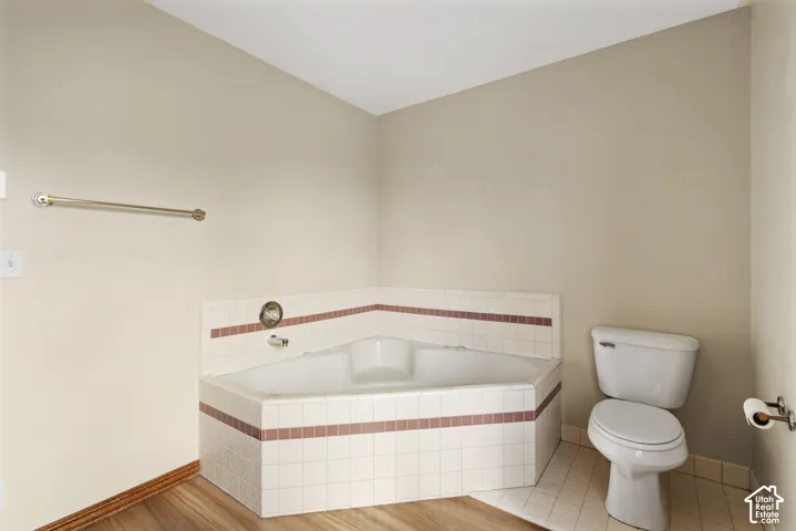 Corner soaking tub with tiled surround, wood-finish flooring, and a commode