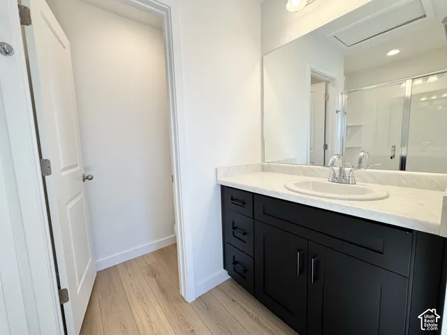 Black vanity with a white countertop and integrated sink, a large wall-mounted mirror, light wood-finish flooring, and a shower with a sliding glass door