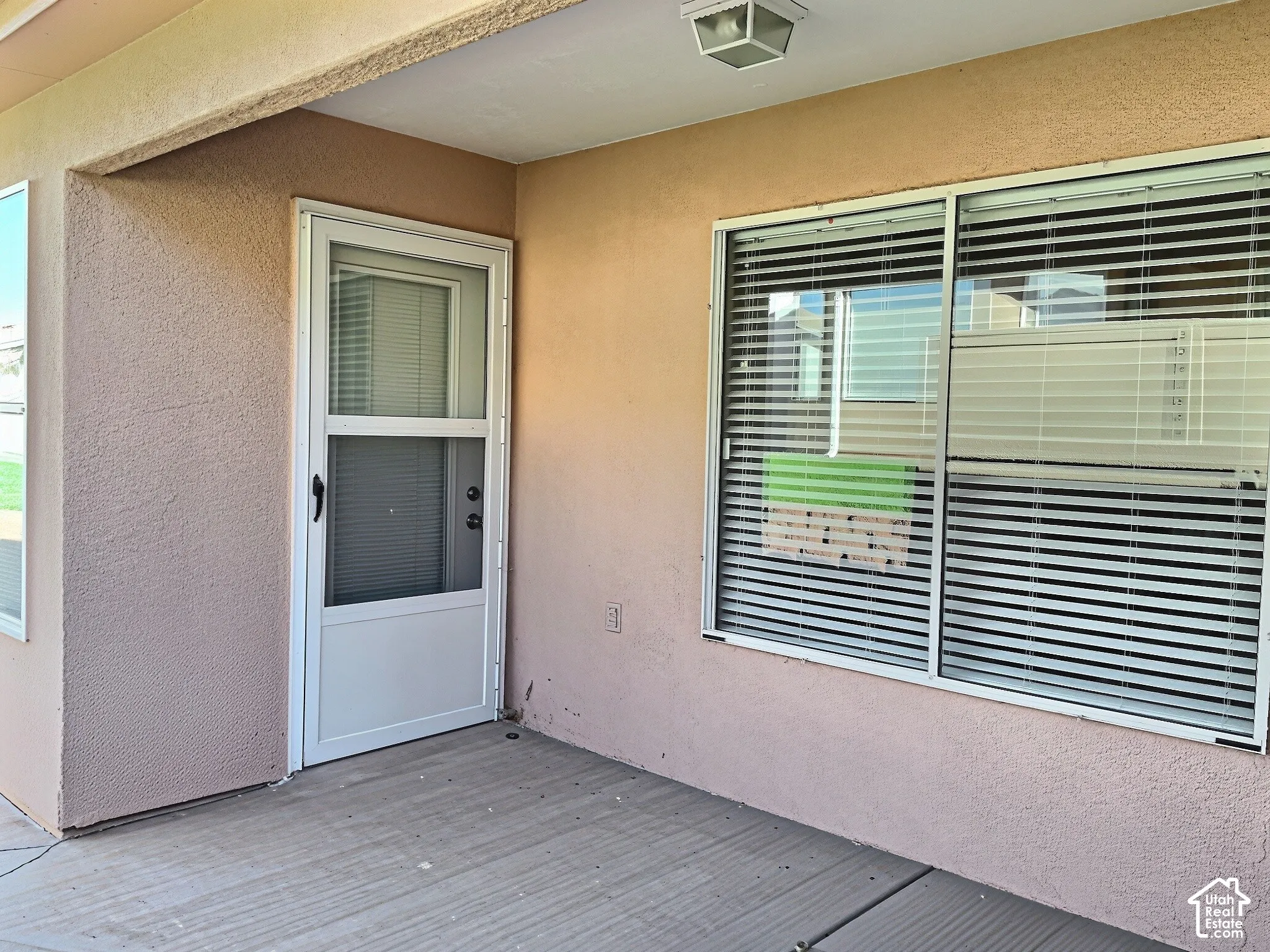 Covered patio area featuring a textured stucco finish, white frame storm door, and sliding windows with horizontal blinds