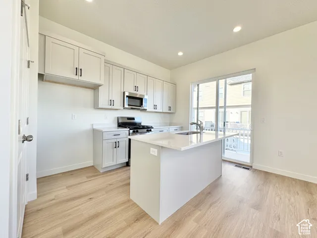 Kitchen featuring white shaker-style cabinetry, stainless steel appliances, a center island with sink, light-toned wood-finish flooring, and a sliding glass door