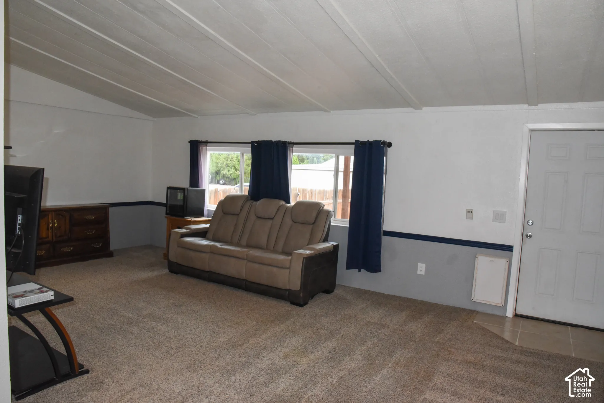 Carpeted living area with two-tone walls, white ceiling with exposed beams, and a white entry door